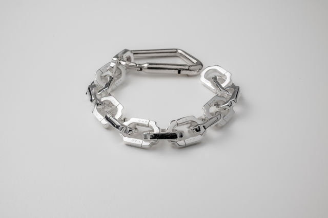 Chunky silver bracelet of faceted flat links, elongated carabiner clasp, laid in a loop on pale grey ground — STRUGA
