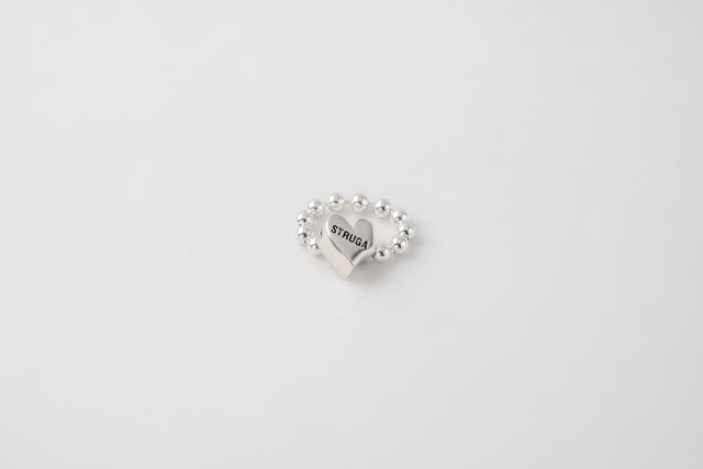 Silver bead ring with a smooth heart at its centre engraved STRUGA, on a pale ground