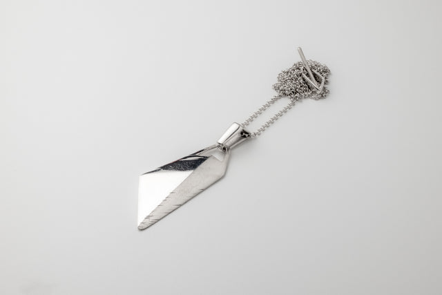Faceted silver blade pendant with filed edge, fine chain coiled beside it, flat lay on pale grey studio surface — STRUGA