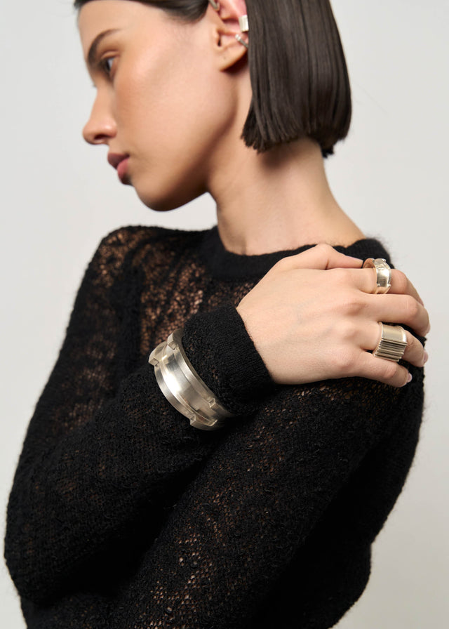 Wide grooved silver cuff worn on a wrist over a black openwork knit sweater, two silver rings on the hand — STRUGA