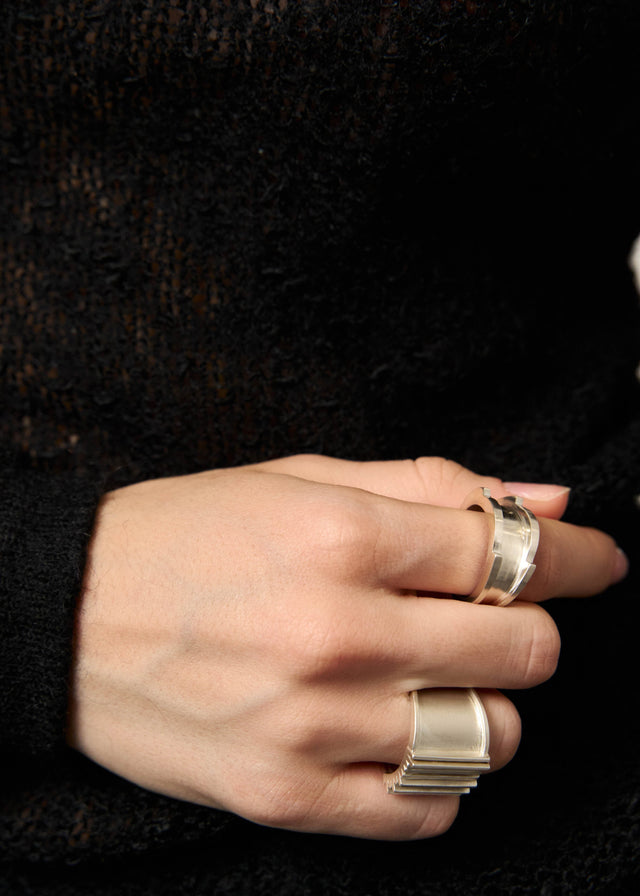 Hand on a black knit sleeve wearing two wide silver rings: a notched band and a ridged stacked band — STRUGA